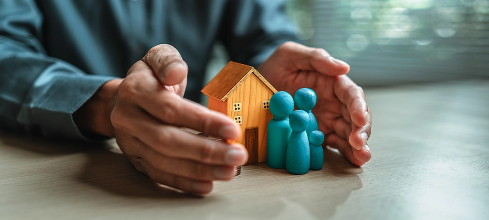Businessman's hand gestures to protect money, house, wooden dolls represent family. Image represents the concept of asset protection, emphasizing safeguarding financial investments and property