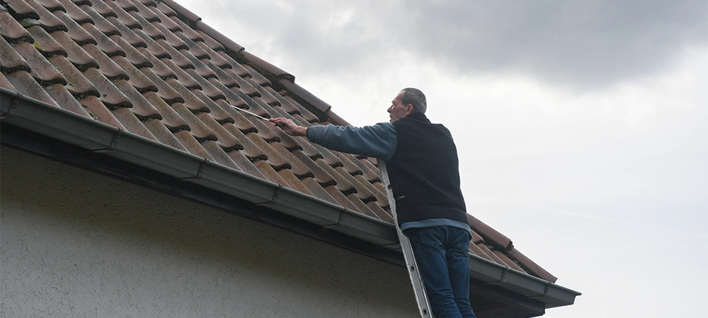 Roofer cleans a gutter with a cleaning tool while standing on a ladder, performing the necessary maintenance to prevent roof leaks and ensure proper drainage of rainwater