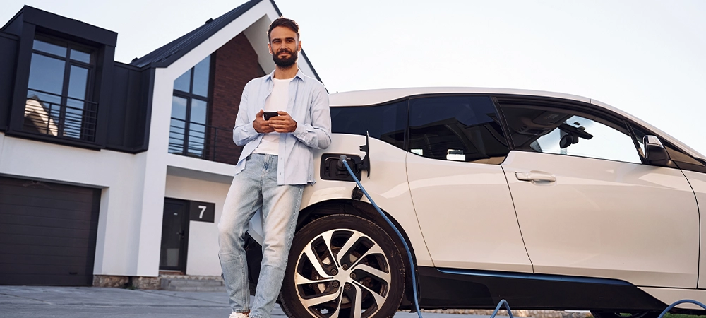 Beautiful young stylish man is with electric car at daytime charging the vehicle and holding smartphone.